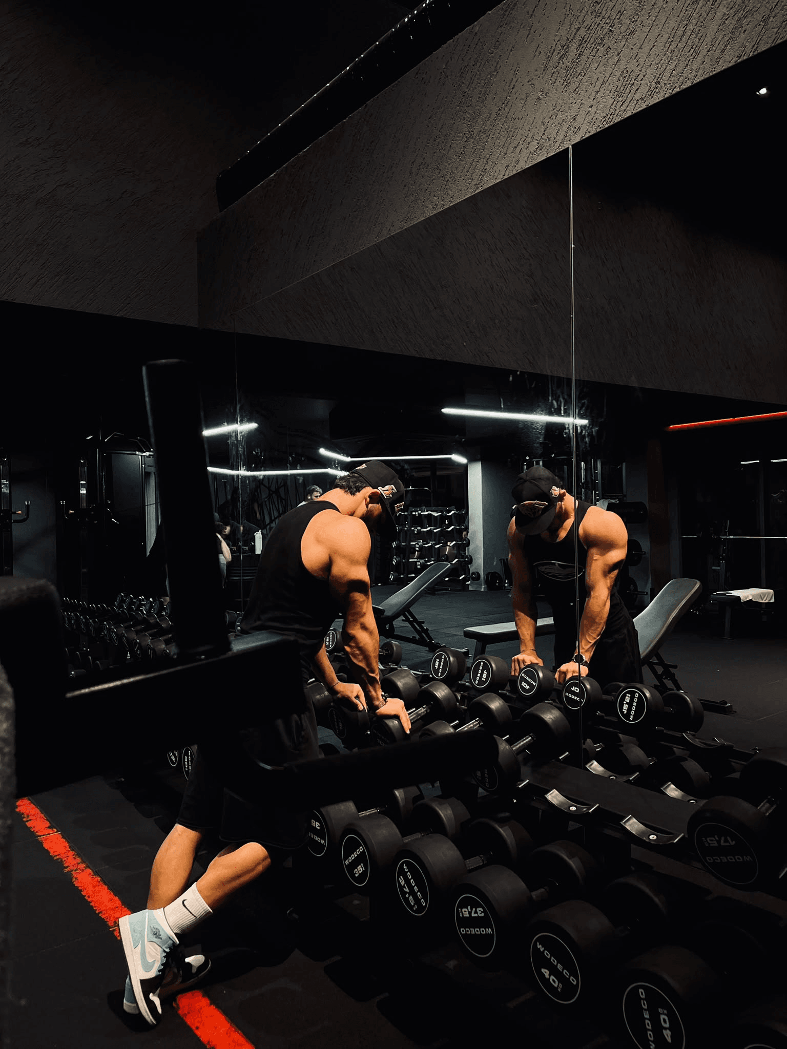 Dambıl rafındaki ağırlıklara uzanan kasketli sporcu; aynada önden yansıması.